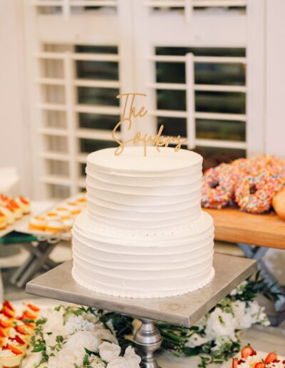 A two-tier white frosted cake with a gold topper, surrounded by floral decor, donuts, and cupcakes on a metal stand.