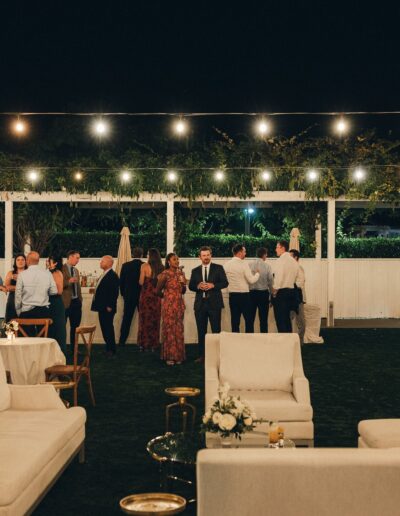 Outdoor gathering at night with string lights, guests mingling, and lounge furniture on a lawn.