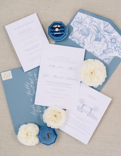 Elegant wedding stationery set with invitations, envelopes, rings in a blue box, and white flowers arranged on a neutral background.