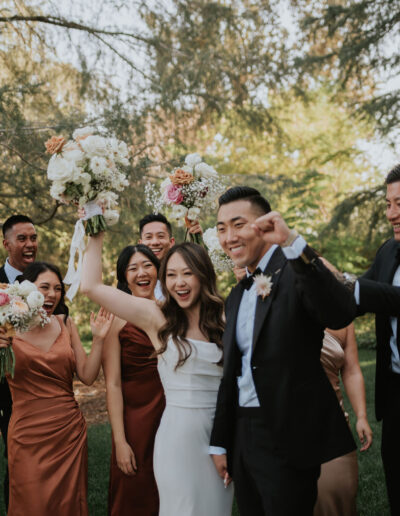 A joyful wedding party celebrating outdoors, with the bride and groom in the center, surrounded by bridesmaids and groomsmen holding bouquets.