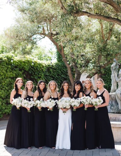 Bride and seven bridesmaids in black dresses pose with bouquets in a garden setting.