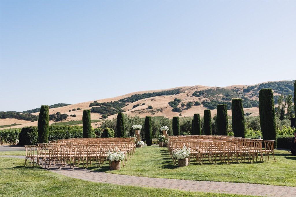 Outdoor wedding setup with rows of wooden chairs, a grassy aisle, and potted plants. Hills and clear blue sky in the background.