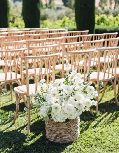 Wicker basket with white flowers placed beside wooden chairs at an outdoor event setting.