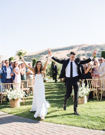 A couple joyfully walks down the aisle outdoors, with guests clapping in the background. The bride holds a bouquet.