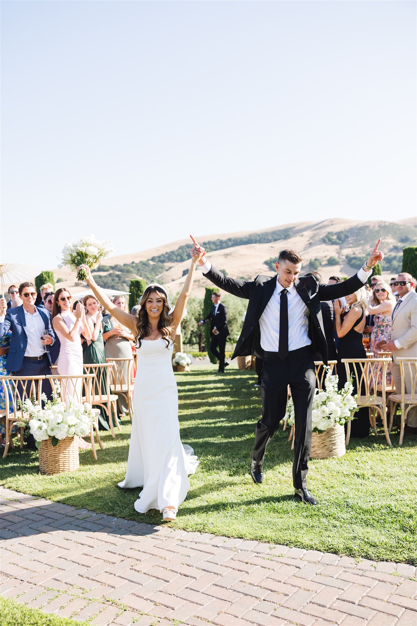 A bride and groom joyfully walk down the aisle outdoors, surrounded by smiling guests seated on either side, on a sunny day.