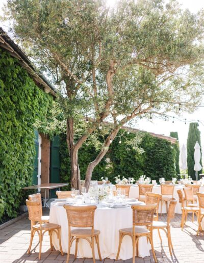 An outdoor dining setup with round tables and rattan chairs under a tree, surrounded by greenery-covered walls.