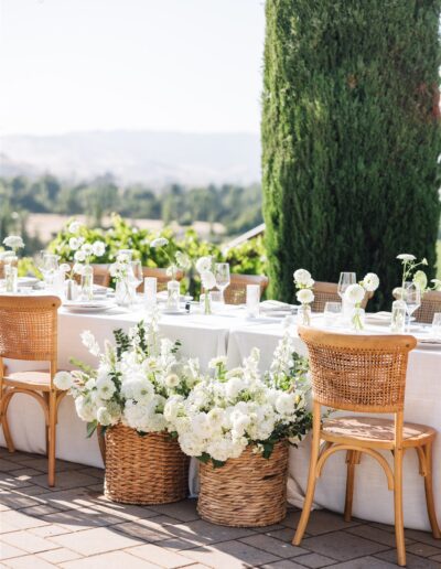 Outdoor dining setup with a long table, wicker baskets of white flowers, wooden chairs, and string lights. A tree and landscape are visible in the background.