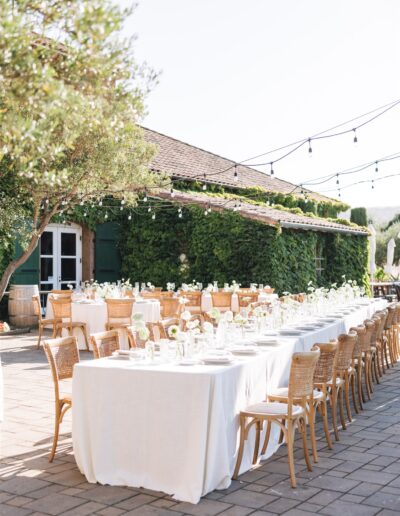 Outdoor dining setup with long tables covered in white tablecloths, wooden chairs, floral centerpieces, and string lights. Greenery and a vine-covered building are in the background.