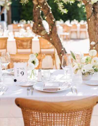 Elegant outdoor dining setup with white tablecloth, floral centerpieces, glassware, and neatly arranged place settings in a sunlit garden.