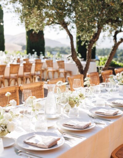 Outdoor dining setup with white tablecloths, plates, glasses, and napkins. Tables are decorated with small white flower arrangements. Trees in the background.