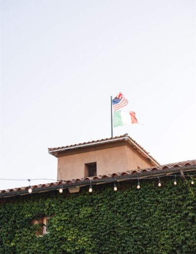 Vine-covered building with string lights and two flags, American and Italian, on the roof against a clear sky.