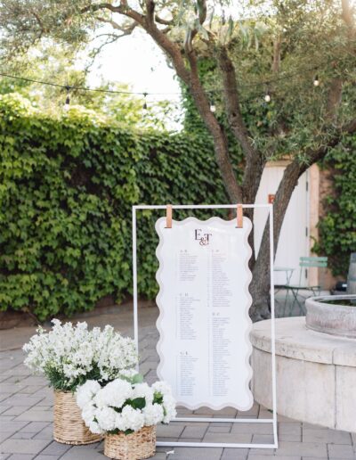 A seating chart on a decorative frame stands outside. Two baskets with white flowers sit beside it on a stone pavement. Greenery and string lights are visible in the background.