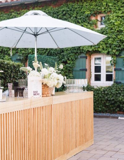 A wooden outdoor bar with a white umbrella, flowers, and glasses, set in front of a brick building covered in green ivy.