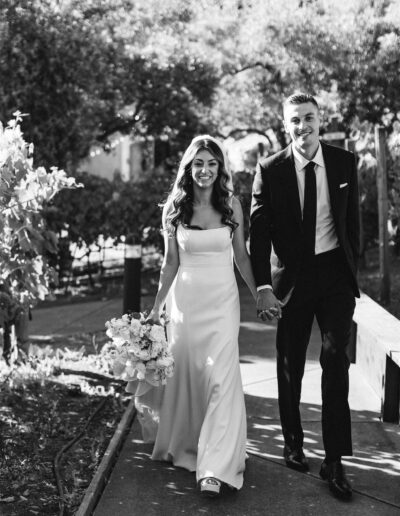 A couple in wedding attire holds hands and smiles while walking outdoors along a path, surrounded by greenery. Black and white photo.