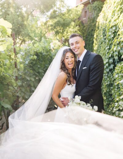 Bride and groom smiling in a garden setting, with the bride wearing a long veil and holding a bouquet.