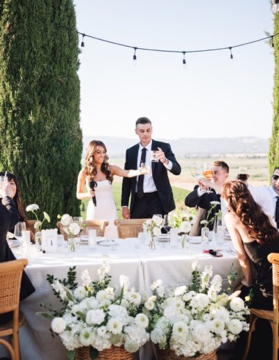 A group of people gather at an outdoor event, holding drinks. The scene features a long table with white flowers and string lights above.