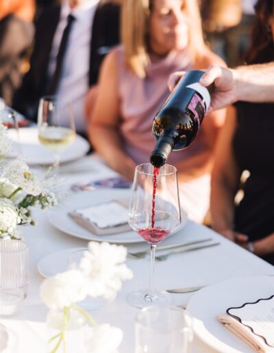 Wine being poured into a glass at a table set for a meal, with people seated in the background.