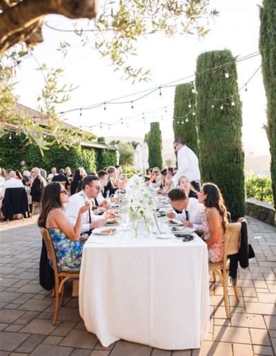 People seated around a long outdoor table, dining under string lights and surrounded by tall trees and greenery, with distant hills visible in the background.