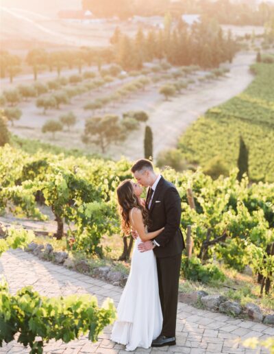 A couple in wedding attire embraces on a stone path in a vineyard at sunset, surrounded by lush green vines and rolling hills.