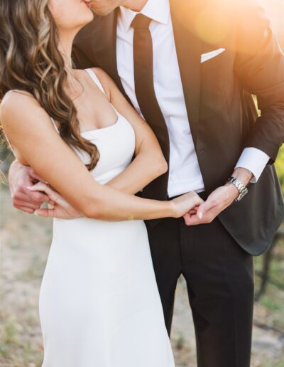 A couple in formal attire shares a kiss, standing closely with the sun shining brightly in the background.