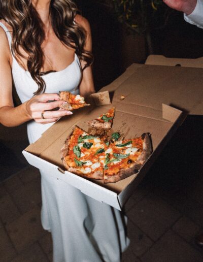 Person in white dress holding a pizza box with a sliced pizza, topped with basil and cheese, while another hand reaches for a slice.