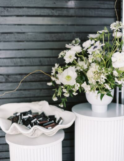 White floral arrangement in a vase on a round pedestal next to a shell-shaped dish holding small rectangular black objects. Background features a dark horizontal wooden slat wall.