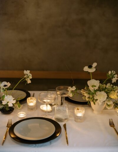 A dining table set with black-rimmed plates, glassware, lit candles, and white floral arrangements on a white tablecloth.