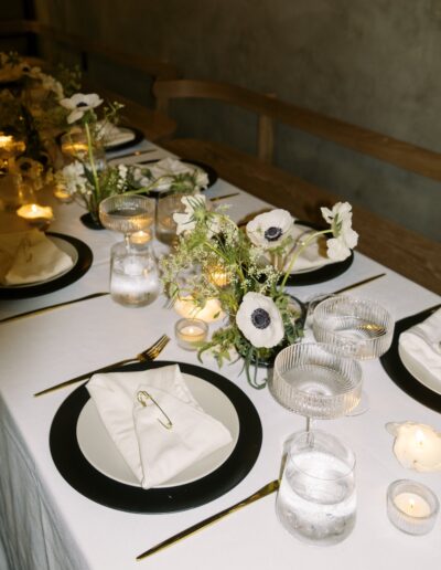 Elegant dining table set with black-rimmed plates, white napkins, glassware, candles, and floral centerpieces featuring white flowers.