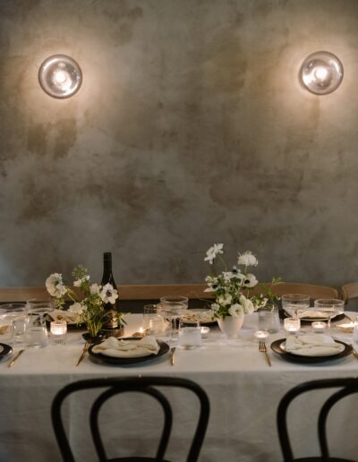 A dimly lit dining table with a white tablecloth, set with plates, glasses, candles, and floral arrangements. Two wall lights illuminate the scene.