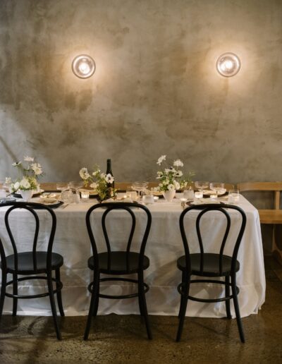 A dining table set for four with a white tablecloth, black chairs, flowers, glassware, and a bottle, against a textured gray wall with two round lights.