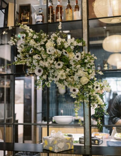 A floral arrangement with white flowers hangs on a grid wall above a table displaying a gift box. Bottles and decor elements are visible in the background.