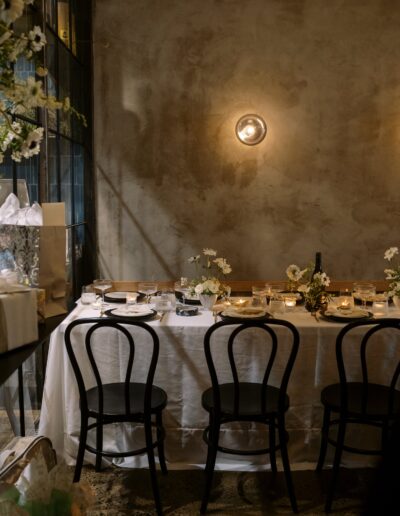 A dimly lit dining area with a table set for a meal, featuring white tablecloth, black chairs, candles, flowers, and a concrete wall in the background.