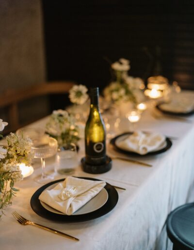 A table set for a formal dinner with a white tablecloth, plates with folded napkins, a bottle of wine, candles, and floral decorations.