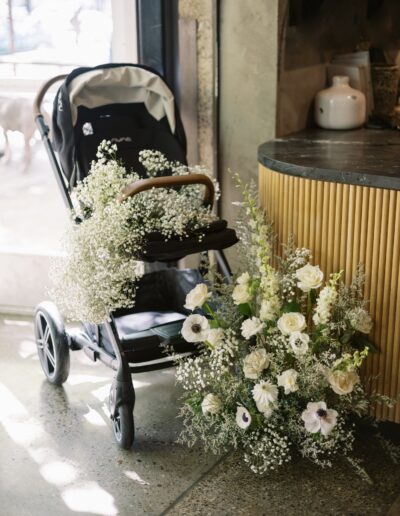 A stroller filled with delicate white flowers is positioned next to a floral arrangement on a countertop inside a room with sunlight streaming in.