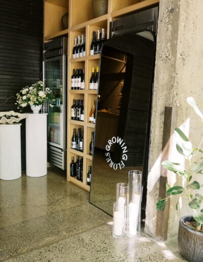 Wine bottles are displayed on wooden shelves next to a mirrored sign reading "GROWING OLD IS GETTING GORGEOUS," with decorative plants and tall candles nearby.