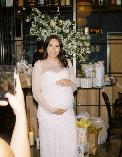 A pregnant woman in a white dress poses for a photo at a celebration, surrounded by flowers and gifts. Another person takes her picture with a phone.