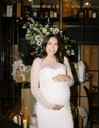 A pregnant woman in a white dress stands smiling in front of a decorated area with flowers and gifts.