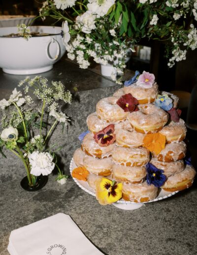 A stack of sugar-coated donuts adorned with edible flowers is displayed on a plate beside a small bouquet and napkins on a concrete countertop.