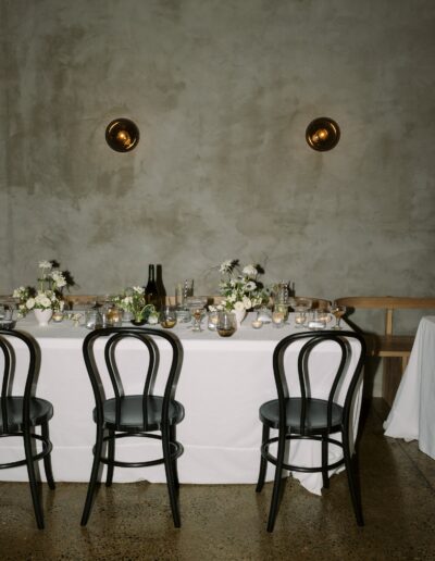 A simple, elegant dining table setup with white tablecloth, black chairs, floral centerpieces, and tableware against a muted gray wall with two round wall lights.