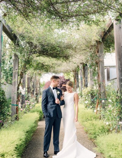 A bride and groom stand together in an outdoor garden walkway, surrounded by greenery and flowers. The groom is in a dark suit, and the bride is in a white gown.