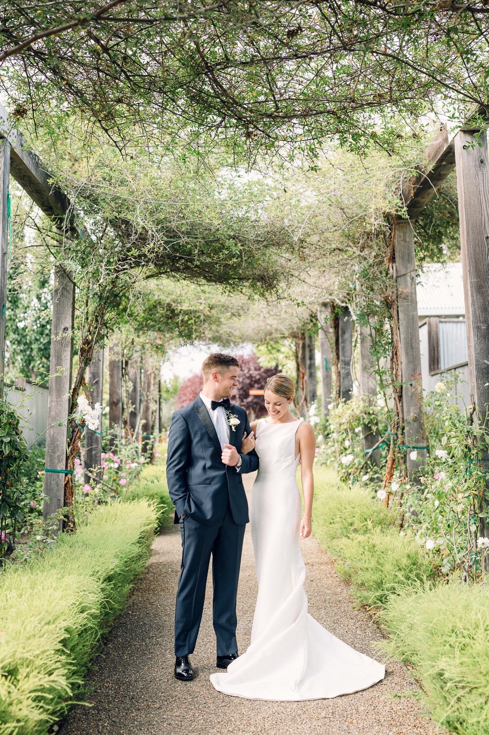 A bride and groom stand together in an outdoor garden walkway, surrounded by greenery and flowers. The groom is in a dark suit, and the bride is in a white gown.