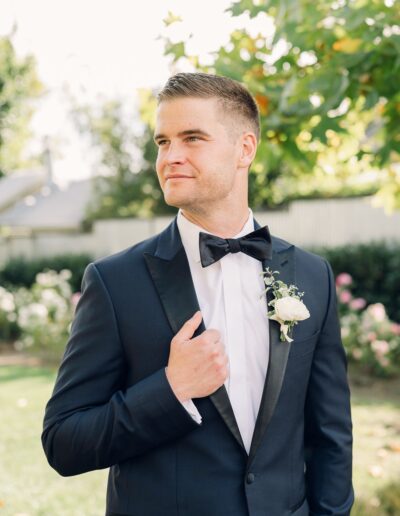 A man in a tuxedo, holding his lapel, stands outdoors with greenery and flowers in the background.