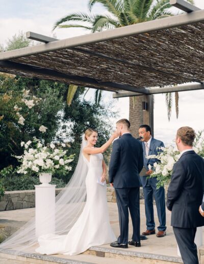 A bride and groom stand at an outdoor altar during their wedding ceremony, with a wedding officiant and a groomsman nearby. Lush greenery and white flowers surround the setting.