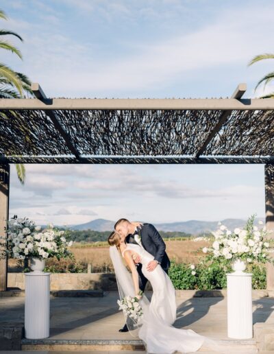 A couple shares a kiss under a pergola during their wedding ceremony, surrounded by floral arrangements and overlooking a scenic landscape.