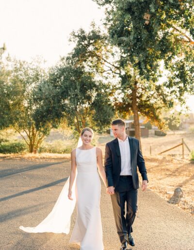 A couple in formal attire walks hand in hand on a sunlit path, surrounded by trees.