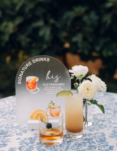 Two cocktails on a floral tablecloth next to a glass vase with white flowers and a sign reading "Signature Drinks: His Old Fashioned.