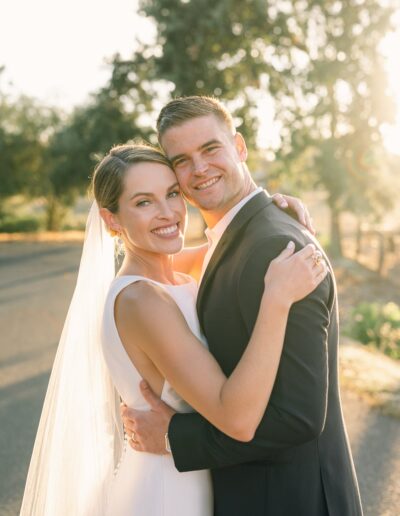 A bride and groom embrace and smile at the camera on a sunlit outdoor path, with trees in the background.