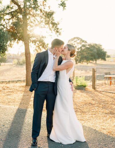 A couple shares a kiss outdoors on a sunny day, dressed in formal attire. The background features trees and a rural landscape.