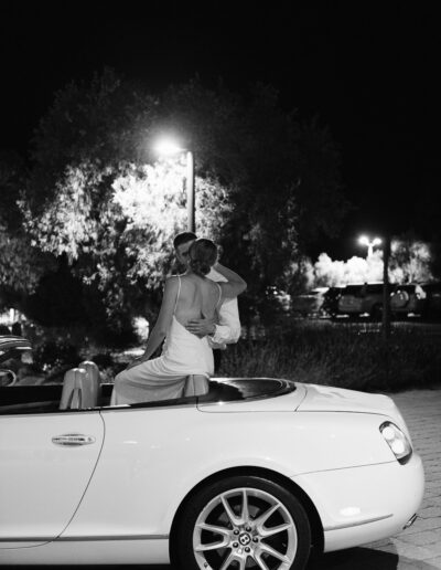 A couple embraces while sitting on the back of a white convertible at night, under streetlights.