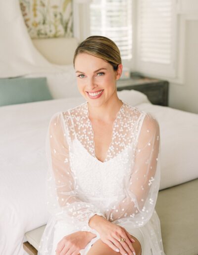 A woman in a white, sheer, embroidered dress sits on a bench in a bright room, smiling at the camera.